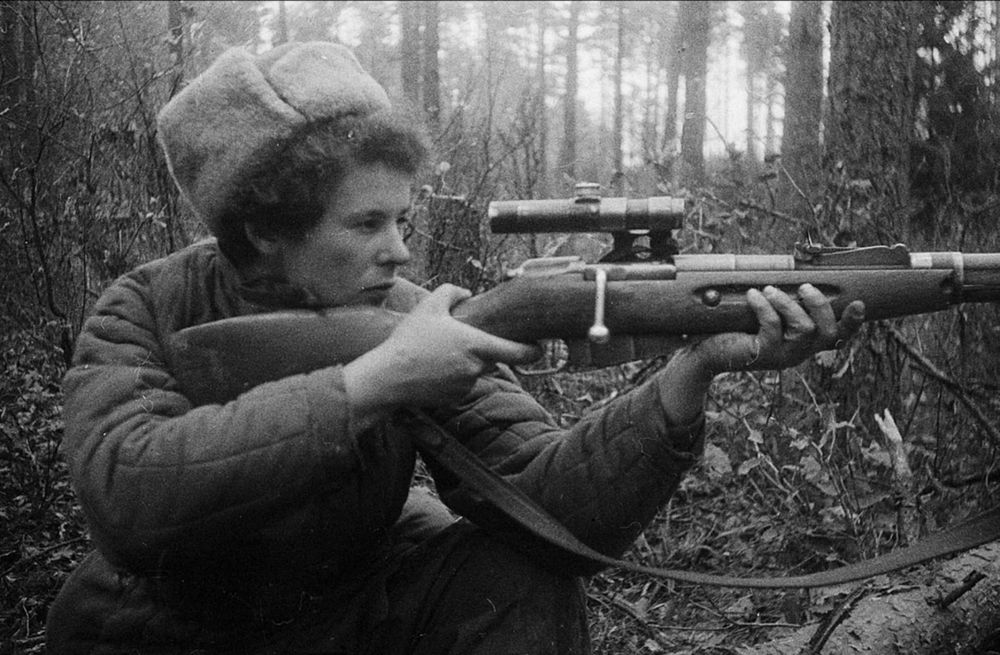 Black-and-white photograph of a woman partisan in a quilted jacket and fur cap, aiming a scoped rifle in a forest